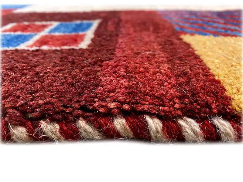 Close-up of a Gabbeh rug: reddish pile with fringes and blue-yellow patterned sections along the edge